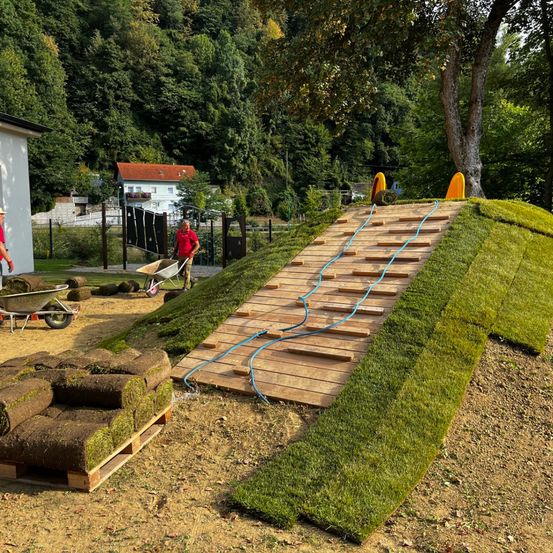 Zwei Personen installieren Holzstufen und grünen Rasen auf einem Hang, in der Nähe eines Baumes und eines Hauses mit rotem Dach. Ein Schubkarren ist mit gerolltem Gras gefüllt.