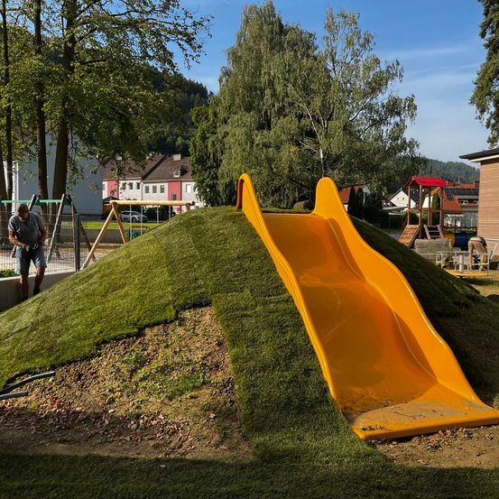 Ein sonniger Tag auf einem Spielplatz mit einer gelben Rutsche, umgeben von Bäumen und einem grasbewachsenen Hügel. Ein Mann steht in der Nähe, und Häuser und Gebäude sind im Hintergrund.