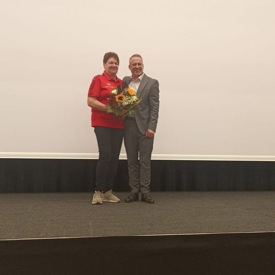 Eine Frau in einem roten Shirt und ein Mann in einem grauen Anzug stehen auf einer Bühne mit einem Blumenstrauß. Sie lächeln beide in die Kamera.