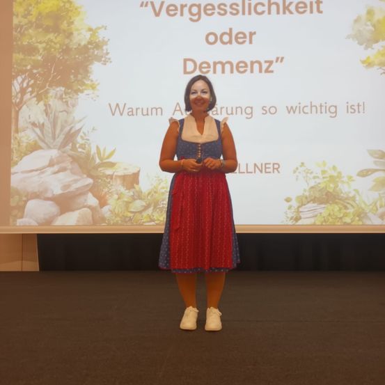 Eine Frau in traditioneller Kleidung steht lächelnd auf der Bühne, vor ihr ein Beamer-Bildschirm mit der Aufschrift 'Vergesslichkeit oder Demenz' und 'Warum Aufklärung so wichtig ist!'.