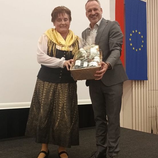 Eine Frau und ein Mann stehen lächelnd zusammen und halten eine Geschenkbox vor einer Europaflagge.