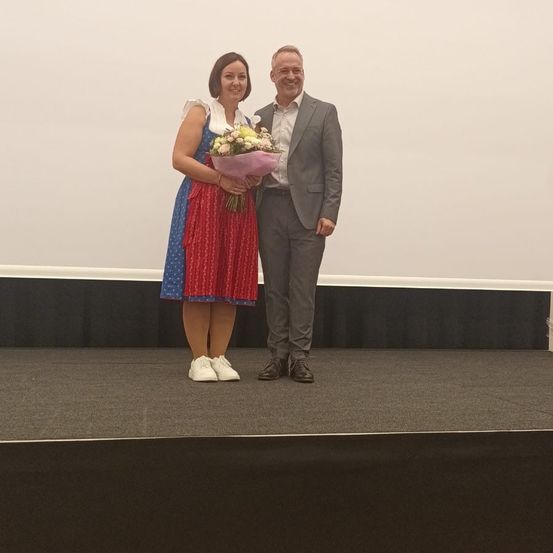 Eine Frau in einem traditionellen Kleid und ein Mann in einem grauen Anzug stehen lächelnd auf der Bühne. Die Frau hält einen Blumenstrauß.