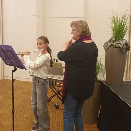 Ein junges Mädchen spielt Flöte, während eine ältere Frau sie begleitet. Beide stehen in einem Raum mit Klavier, Pflanzen und Holzboden.