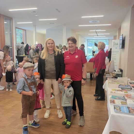 Eine Gruppe von Menschen, darunter Frauen und Kinder, steht in einem Flur mit einer Frau in einem roten Shirt, die lächelt. Einige Kinder tragen traditionelle Kleidung. Ein Tisch mit Büchern steht hinter ihnen.