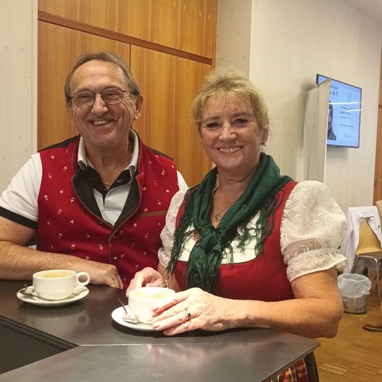 Ein älterer Mann und eine Frau sitzen lächelnd an einer Theke. Der Mann trägt ein rotes Weste, und die Frau ein grünes Tuch. Sie halten beide Tassen mit Untertassen. Ein Fernseher und ein Mülleimer stehen hinter ihnen.