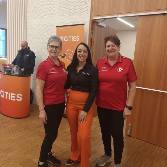 Drei Frauen in roten Uniformen stehen nebeneinander in einem Konferenzraum. Hinter ihnen arbeitet ein Mann an einem Laptop. Ein Banner mit dem Wort 'cities' ist im Hintergrund zu sehen.