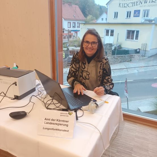 Eine Frau sitzt an einem Schreibtisch mit einem Laptop, einem Schild für Lungenfunktionstests und einem Drucker. Sie lächelt und trägt eine Brille.
