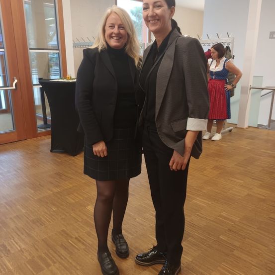 Zwei Frauen stehen lächelnd in einem Raum mit Holzboden nebeneinander. Sie sind in formeller Kleidung gekleidet. Hinter ihnen steht eine Frau in traditioneller Kleidung in der Nähe eines Tisches.