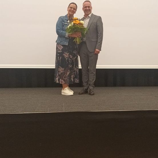 Ein lächelnder Mann und eine Frau stehen mit Blumen auf der Bühne. Die Frau trägt ein Blumenkleid und Turnschuhe, der Mann einen Anzug. Sie stehen sich gegenüber, der Hintergrund ist eine schlichte weiße Wand.