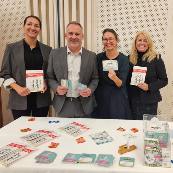 Vier Personen stehen hinter einem Tisch mit verschiedenen Werbematerialien, darunter Broschüren, Karten und Figuren. Der Mann in der Mitte hält eine Karte und eine Broschüre, während die Frauen auf beiden Seiten ebenfalls Karten und Broschüren halten. Der Tisch ist mit einem weißen Tuch bedeckt und hat zusätzliche Karten und Figuren.