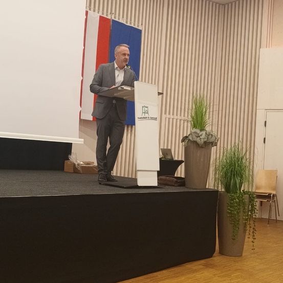 Ein Mann steht auf einer Bühne mit einem Podium und einem Mikrofon. Hinter ihm sind zwei Flaggen und ein Topf mit Pflanzen. Auf der rechten Seite befindet sich ein Stuhl. Die Bühne hat einen schwarzen Boden und eine weiße Wand.