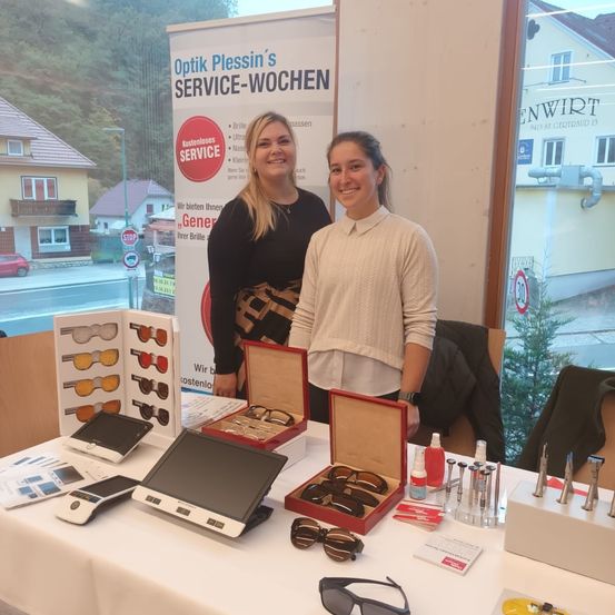 Zwei Frauen stehen hinter einem Tisch mit diverser Brillen, einem Monitor und anderen Gegenständen. Ein Banner hinter ihnen lautet 'Optik Plessin's Service-Wochen'.