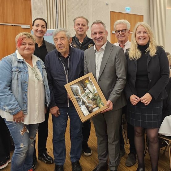 Mehrere Menschen stehen drinnen, lächeln und posieren für ein Foto. Ein Mann im Anzug hält eine gerahmte Box, während eine Frau neben ihm die Hände zusammenhält. Hinter ihnen trägt ein anderer Mann einen Fliegenbinder. Im Hintergrund befindet sich eine Holztür und eine Wand mit einem Schild.