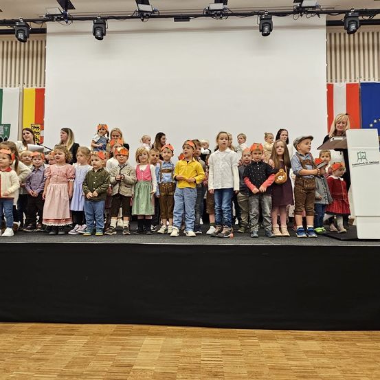 Eine Gruppe von Kindern, die Mützen und Schuhe tragen, steht auf der Bühne und sieht das Publikum an. Eine Frau steht hinter einem Rednerpult mit Fahnen im Hintergrund.