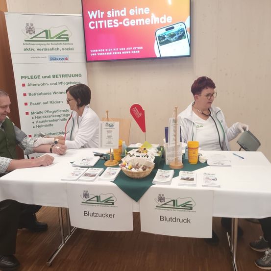 Drei Personen sitzen an einem Tisch mit einer weißen Tischdecke und führen anscheinend ein gesundheitsbezogenes Gespräch. Hinter ihnen steht ein Banner mit Pflege und Betreuung. Eine Projektionsleinwand zeigt eine Werbung für eine Gemeinde namens CITIES.