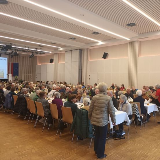Ein großer Raum mit vielen Menschen an Tischen, einer Präsentationsleinwand an der Wand und einer Frau, die in der Nähe der Tische steht.