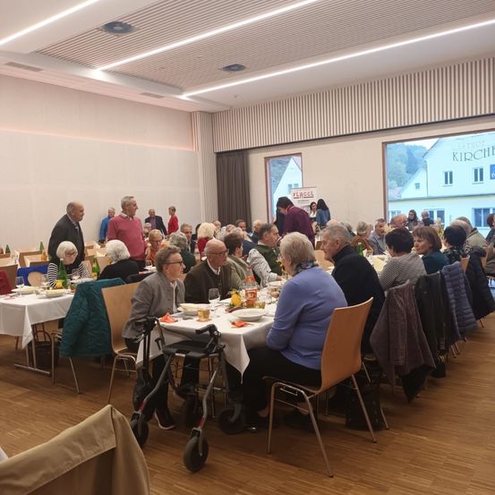 Eine Gruppe älterer Menschen sitzt an Tischen in einem Speisesaal, einige stehen. Der Saal hat große Fenster, einen Fliesenboden und Stühle mit Jacken. Außerhalb der Fenster sind Gebäude zu sehen.