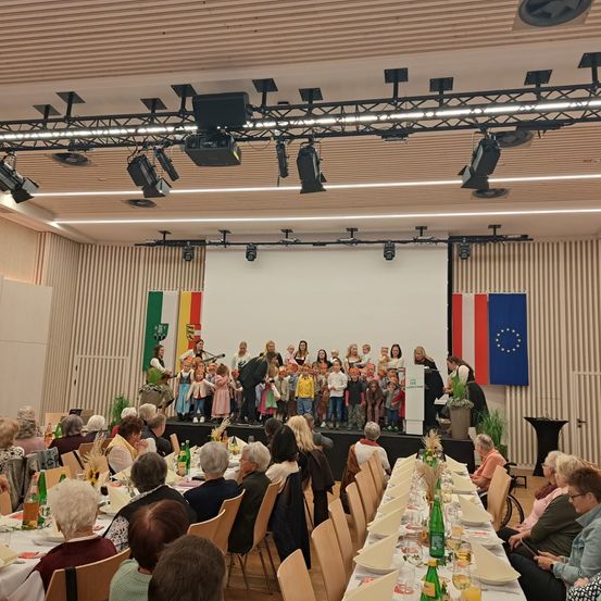 Eine Veranstaltung in einem Saal mit Bühne, Flaggen und einer Menschenmenge älterer Menschen. Die Bühne hat Musikinstrumente und Kinder in traditionellen Kostümen. Der Raum ist gut beleuchtet mit Deckenleuchten.