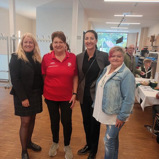 Vier Frauen stehen zusammen in einem Raum mit Holzböden. Die Frau in der Mitte trägt ein rotes Shirt mit einem Logo, während die anderen lässig gekleidet sind. Dahinter stehen Tische mit weißen Tischdecken, an denen Leute sitzen.