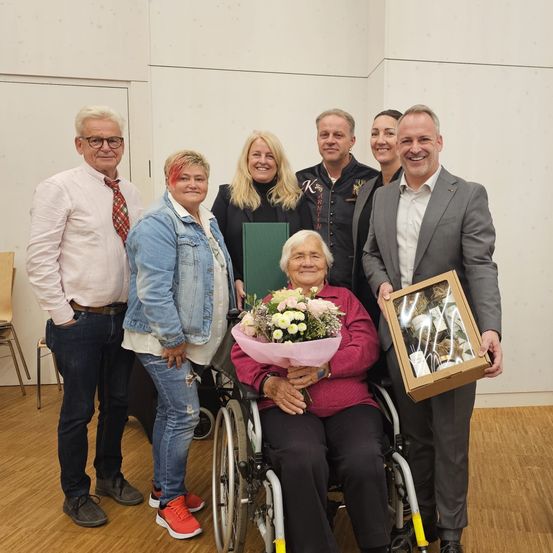 Eine Gruppe von Menschen steht in einem Raum mit einer Frau im Rollstuhl, die Blumen und einen Strauß hält. Ein Mann neben ihr hält eine gerahmte Schachtel. Sie alle lächeln und scheinen ein Ereignis zu feiern.