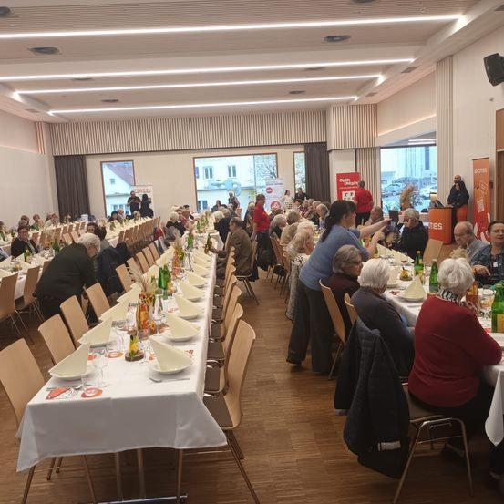 Eine große Gruppe älterer Menschen sitzt an langen Tischen in einem Speisesaal. Die Tische sind mit Tellern, Gläsern und Flaschen gedeckt. Einige Leute stehen und unterhalten sich. Der Raum hat große Fenster und einen an der Wand montierten Lautsprecher.