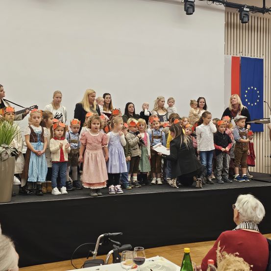 Kinder in traditionellen Kostümen treten auf der Bühne auf, begleitet von einer Frau mit Mikrofon und einer Frau, die Gitarre spielt. Im Hintergrund ist eine Flagge zu sehen.