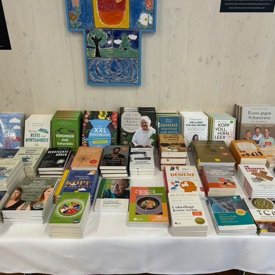 Ein weißer Tisch zeigt eine Sammlung von Büchern. Titel umfassen 'Demenz verstehen und Mutig leben', 'XXL Kochbuch' und 'Organisiert'. Ein farbenfrohes Gemälde hängt an der Wand dahinter.