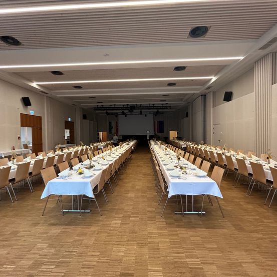Ein leerer Bankettsaal mit langen Tischen und Stühlen. Im Hintergrund befindet sich eine Projektionswand. Der Raum hat einen Holzboden und weiße Wände.