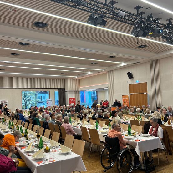 Ein großer Raum mit Holzboden ist mit Menschen an langen Tischen gefüllt. Flaschen, Teller und Servietten sind auf den Tischen. Die Decke des Raums hat Lichter und Lautsprecher. Eine Person im Rollstuhl ist unter den Teilnehmern.