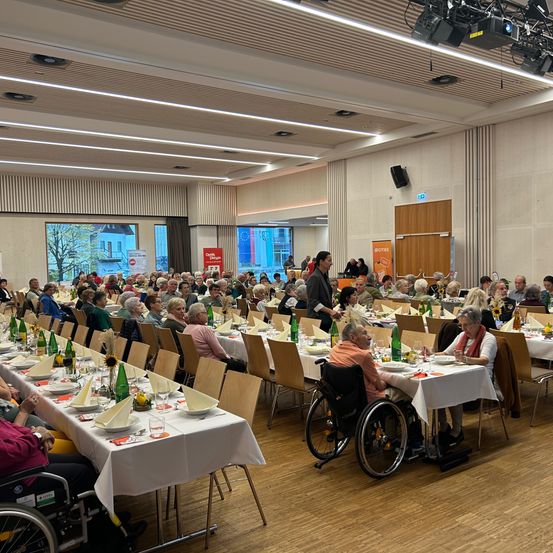 Ein großer Saal mit mehreren langen Tischen, an denen Menschen sitzen und essen. Auf den Tischen stehen viele grüne Flaschen, Teller und Servietten. Eine Frau steht und spricht mit den Menschen. Einige Menschen sind im Rollstuhl. Der Saal hat Holzböden, Deckenleuchten und eine Glaswand.