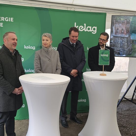 Vier Personen stehen hinter weißen Podesten, lächeln und schauen in die Kamera. Hinter ihnen befindet sich ein grüner Banner mit der Aufschrift 'kelag'. Im Hintergrund zeigt ein großes Bild eine Burg.