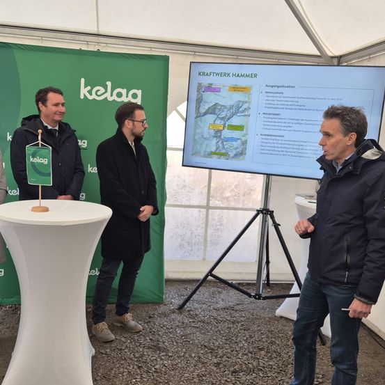 Drei Männer stehen in einem Zelt. Ein Mann spricht, ein anderer hört zu. Eine Leinwand zeigt eine Präsentation über Kraftwerk Hammer.