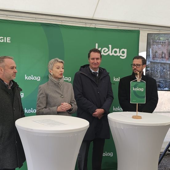 Vier Personen stehen vor einem grünen Banner mit dem Wort 'kelag' in weiß. Sie lächeln und scheinen für ein Foto zu posieren. Der Mann rechts hält ein grünes Schild mit dem Wort 'kelag'.