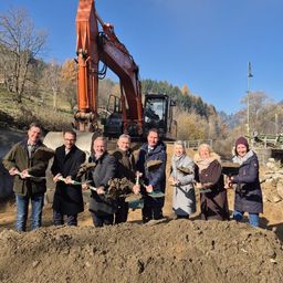 Eine Gruppe von Menschen in Winterkleidung posiert mit Schaufeln in einem Erdhaufen. Hinter ihnen steht ein großer oranger Bagger. Bäume und ein blauer Himmel sind im Hintergrund.
