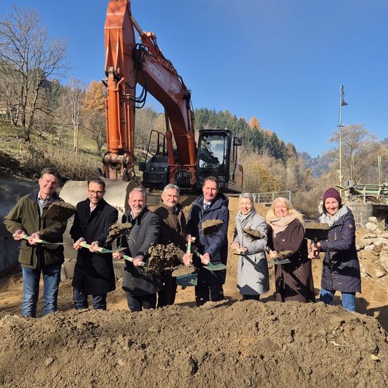 Eine Gruppe von Menschen in Winterkleidung posiert mit Schaufeln in einem Erdhaufen. Hinter ihnen steht ein großer oranger Bagger. Bäume und ein blauer Himmel sind im Hintergrund.