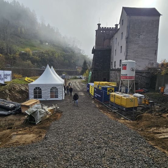 Eine Baustelle mit Schotterweg und Zelten in einem Gebirgsgebiet. Mehrere Menschen gehen herum. Im Hintergrund steht ein Gebäude.