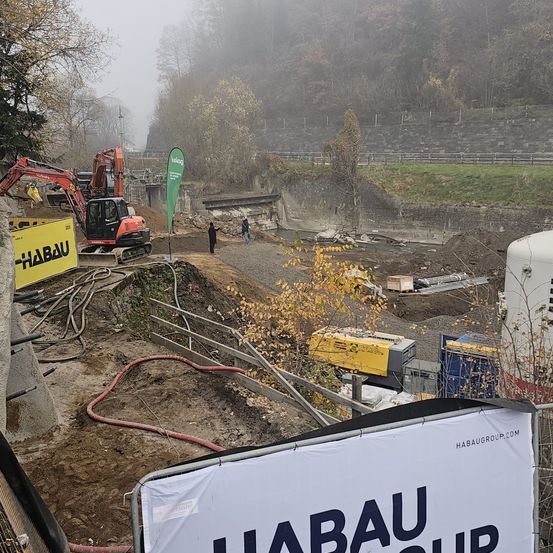Eine Baustelle mit schweren Maschinen, Arbeitern und Bannern. Bäume und Pflanzen sind in der Umgebung. Ein Fluss fließt in der Nähe mit einer Brücke im Hintergrund.