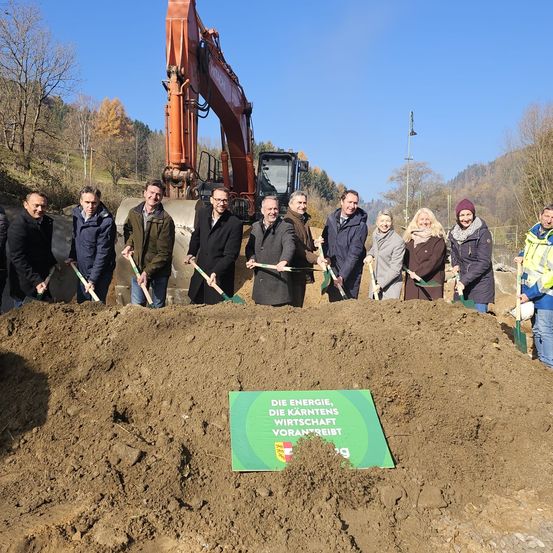 Eine Gruppe von Menschen, möglicherweise Beamte, steht in einem Haufen Erde. Sie halten Schaufeln, möglicherweise für eine feierliche Grundsteinlegung. Ein grünes Schild befindet sich in der Mitte der Erde. Ein großer Bagger steht hinter ihnen. Bäume und Berge sind im Hintergrund.