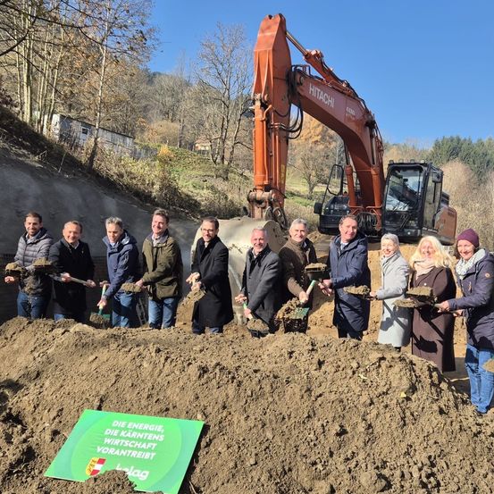 Eine Gruppe von Menschen in Winterkleidung, die Schaufeln halten, posiert für ein Foto an einer Baustelle mit einem großen orangefarbenen Bagger im Hintergrund. Ein grünes Schild steht 'Die Energie, die Kärntens Wirtschaft vorantreibt'.