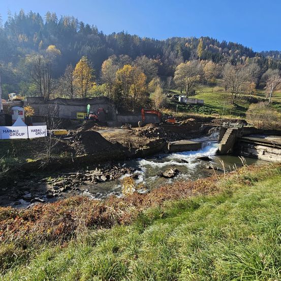 Ein Baugelände mit Baggern und einem Schild mit der Aufschrift 'HABAU GROUP' befindet sich an einem Fluss. Der Fluss fließt durch Felsen, und die Umgebung ist mit Bäumen und Bergen gefüllt.