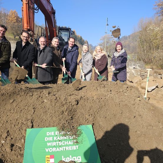 Eine Gruppe von Menschen in Winterkleidung posiert mit Schaufeln an einer Baustelle, ein Schild mit der Aufschrift 'Die Erste - Die Karntner Wirtschaftsvorantritt' ist zu sehen. Hinter ihnen ist ein Bagger zu sehen, und die Szene spielt in einer natürlichen Umgebung mit Bäumen und Bergen.