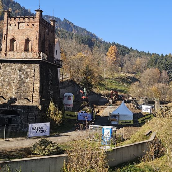 Eine Baustelle befindet sich in der Nähe eines alten Backsteingebäudes. Die Baustelle weist ein Zelt, Banner und Baumaschinen auf. Im Hintergrund befindet sich ein mit Bäumen und Pflanzen bedeckter Hügel.