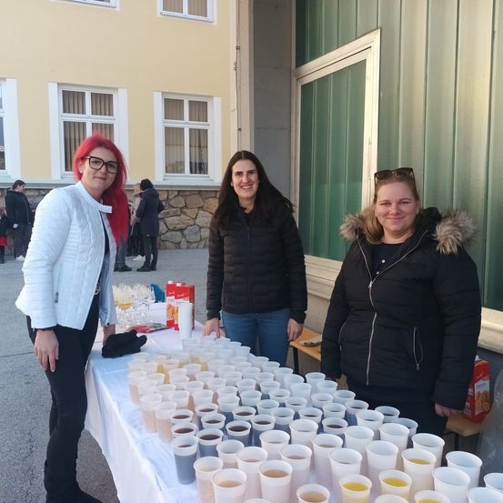 Drei Frauen stehen hinter einem Tisch mit gefüllten Getränkebechern und lächeln für ein Foto vor einem Gebäude.
