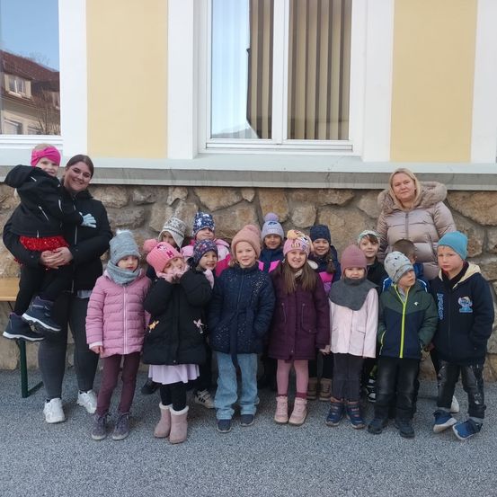 Eine Gruppe von Kindern und zwei Frauen steht vor einem Steingebäude mit einem Fenster und weißen Rahmen. Sie tragen Winterkleidung und einige haben Mützen auf.