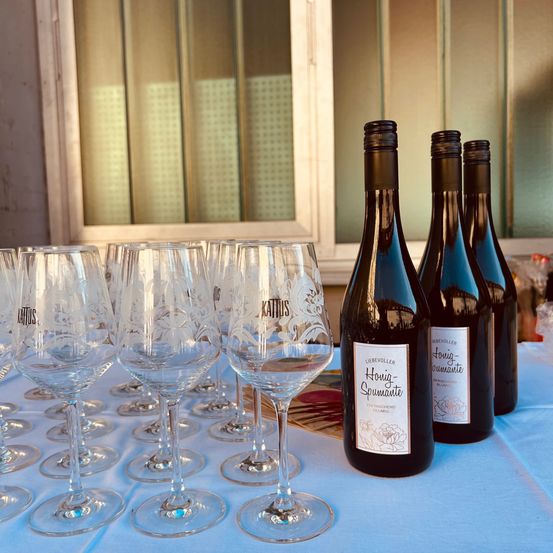 Ein Tisch mit Weingläsern und Flaschen von Liebvoll Honig-Spumante. Die Umgebung ist in der Nähe eines Fensters mit Blick auf einen Garten.