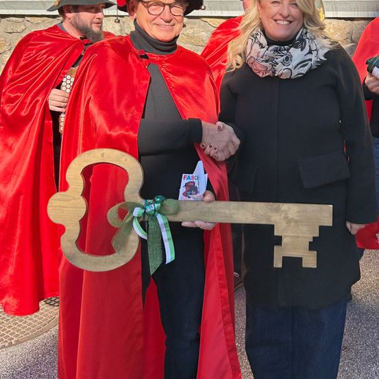 Ein Mann in einem roten Umhang und eine Frau in einem schwarzen Mantel halten einen großen hölzernen Schlüssel und lächeln. Hinter ihnen stehen zwei Personen in roten Umhängen, eine hält eine Flasche.