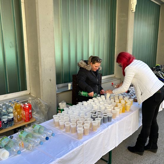 Zwei Frauen stehen bei einem Tisch voller Plastikbecher und bereiten Getränke zu. Eine hält eine Flasche und die andere einen Becher. Hinter ihnen sind zwei Fenster und Flaschen mit Wasser und Saft.
