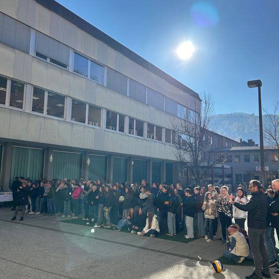 Eine große Gruppe von Menschen steht und sitzt vor einem Gebäude, mit einem Mann, der kniet und einen Ball hält, wahrscheinlich bei einer Schulveranstaltung unter klarem Himmel.