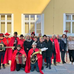 Eine Gruppe von Menschen in roten Kostümen und Hüten posiert für ein Foto vor einem Gebäude. Einige halten Musikinstrumente und Requisiten.