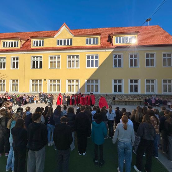 Eine Menschenmenge versammelt sich vor einem gelben Gebäude mit rotem Dach. Einige Personen tragen rote Umhänge, während andere Jacken und Turnschuhe tragen. Das Gebäude hat mehrere Fenster und einen kleinen Balkon.